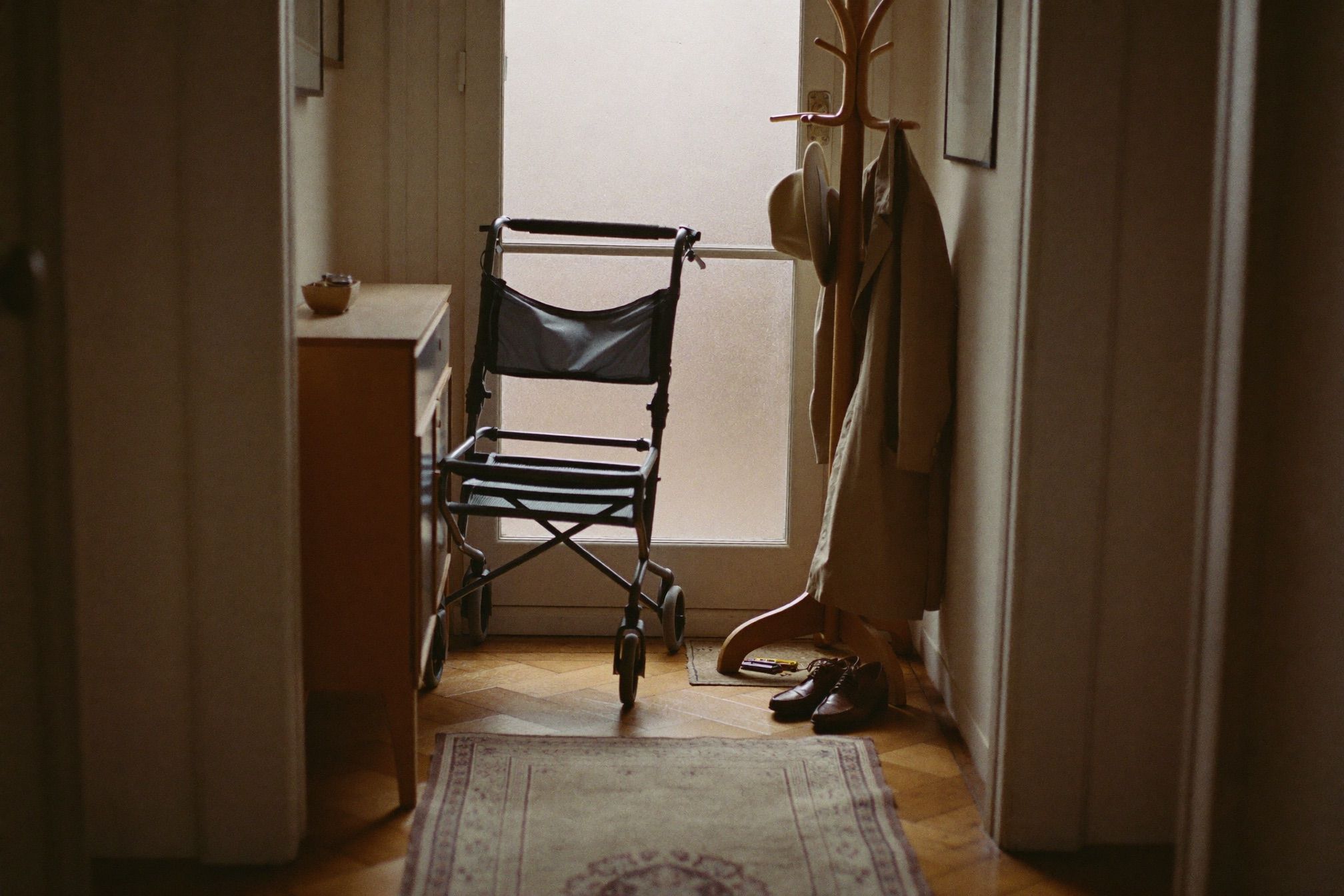 Ein Rollator im Flur einer Altbauwohnung neben einer Garderobe mit Mantel und Hut, Morgenlicht durch die Milchglastür