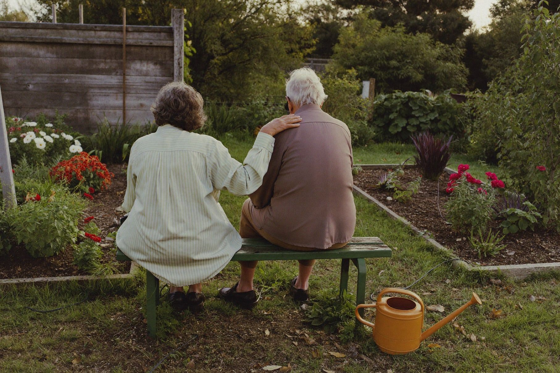 Älteres Paar von hinten auf einer Gartenbank im Schrebergarten, daneben eine Gießkanne